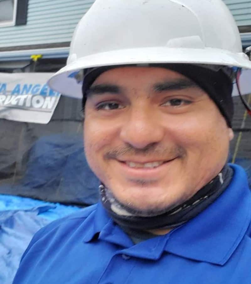 A construction worker wearing a hard hat and a blue shirt, smiling for a selfie at a job site, specializing in storm damage roofing.